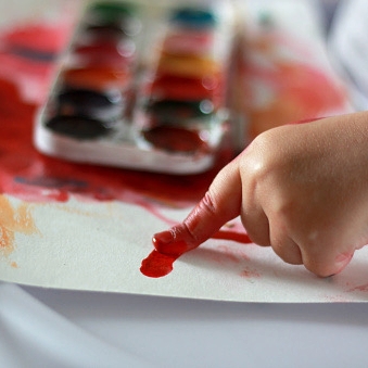Photo child draws a finger in red paint on paper. hands in paint. against a watercolor paint background. children's creativity, painting, early development, fine motor skills